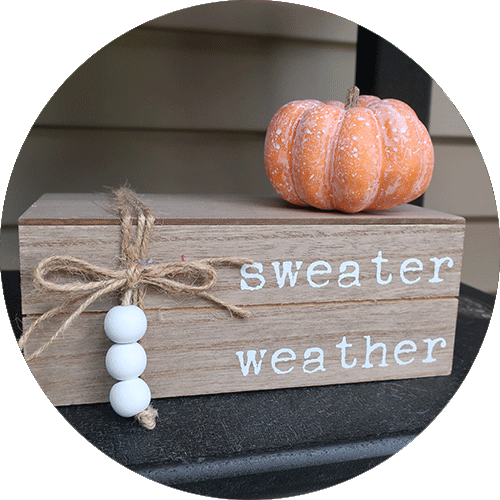 A wooden block that says sweater weather, with an orange pumpkin on top.